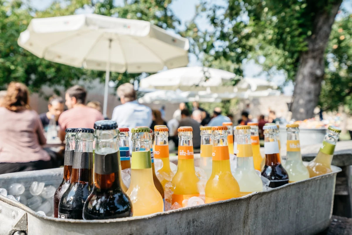 Verschiedene Bierflaschen in unterschiedlichen Farben und Größen in einer Metallwanne, im Hintergrund ein weißer Sonnenschirm und verschwommene Personen auf einer Sommerparty.