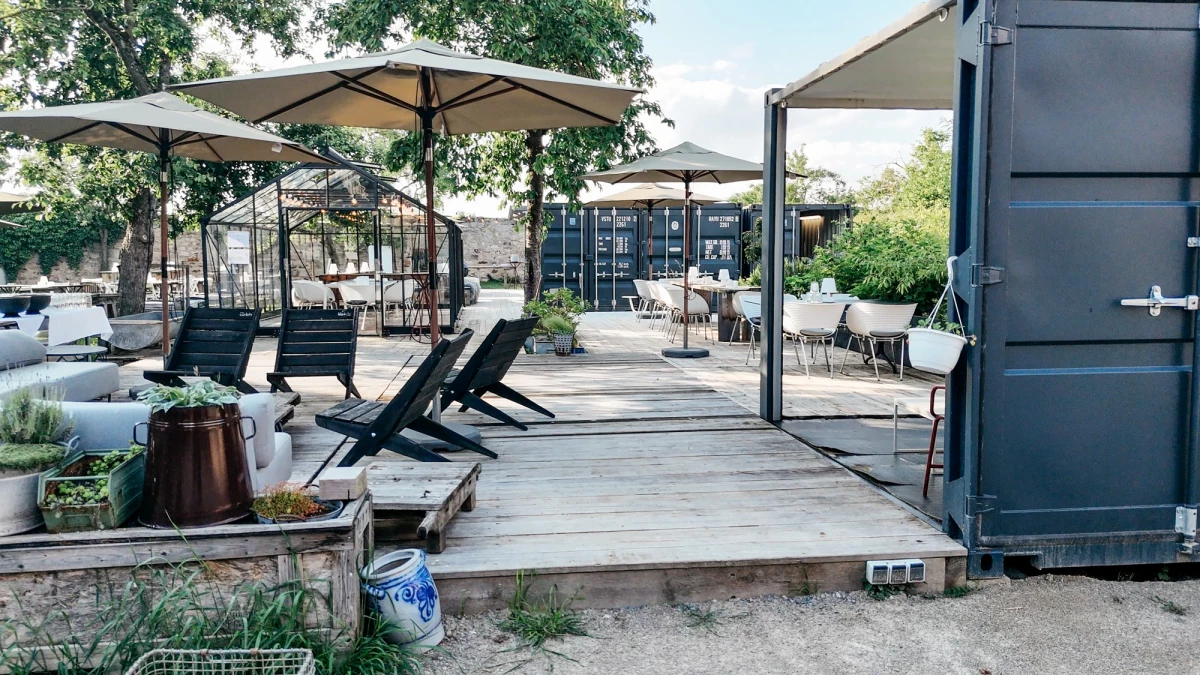 Moderne Terrasse mit grauen Liegen unter Sonnenschirmen, holzgepflasterte Fläche mit Sitzgelegenheiten und einem grauen Container im Seitenbereich, umgeben von Bäumen und Pflanzen.