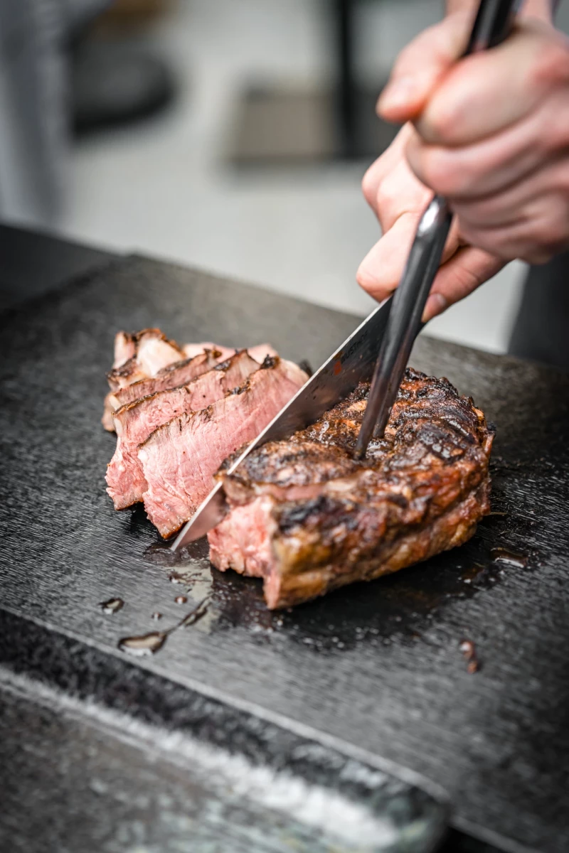 Gebratenes, rosafarbenes Rindfleisch wird mit einer Grillzange auf einer dunklen Steinplatte geschnitten. Das saftige Steak ist medium gegart und hat eine appetitliche, angeröstete Kruste.