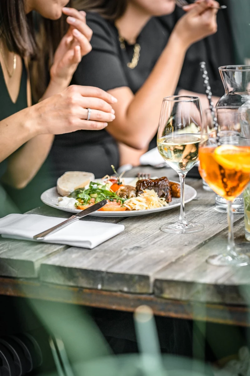 Gedeckter Holztisch mit einem Teller mit Hauptgericht, Salat und Besteck, daneben ein Glas Weißwein und ein orangefarbener Cocktail. Personen in dunkler Kleidung sitzen am Tisch und greifen nach Essen und Gläsern.
