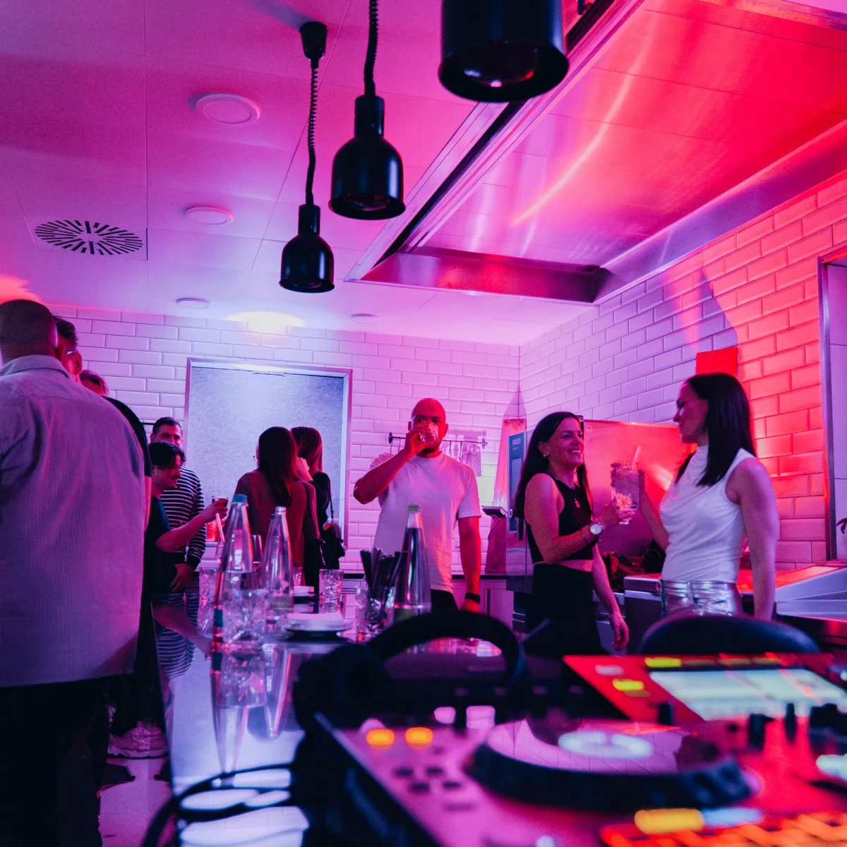 Personen in einer Bar mit dramatischer rot-pinker Beleuchtung, Hängelampen und Ziegelwänden. Auf dem Tresen stehen Gläser und Flaschen, im Vordergrund ist ein DJ-Pult zu sehen. Die Atmosphäre ist urban und club-ähnlich.