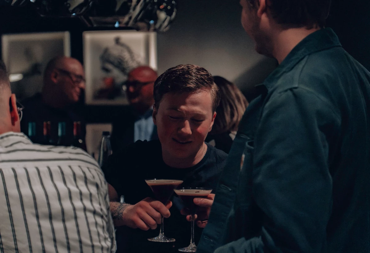 Eine Gruppe von Männern an einer Bar, die einen Drink genießen. Einer hält ein Cocktailglas mit einem dunklen Getränk und trägt ein gestreiftes Hemd. Die Atmosphäre ist gedämpft und intim.