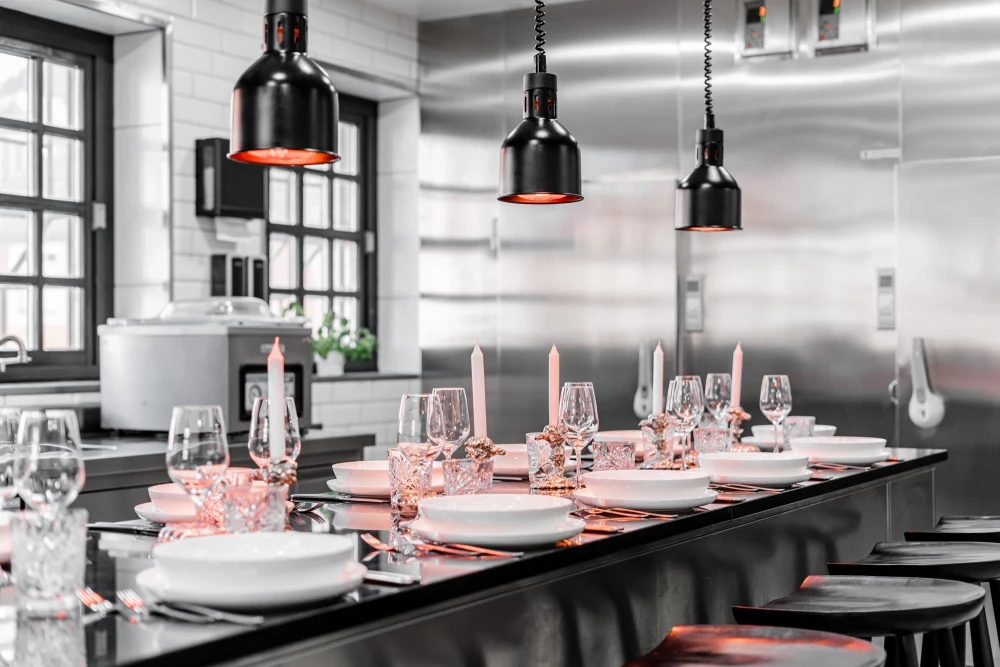 Gedeckter Tisch für ein Abendessen mit weißen Tellern und Weingläsern unter schwarzen Pendelleuchten in einer modernen Edelstahl-Küche mit Theke und Industriedesign-Atmosphäre.