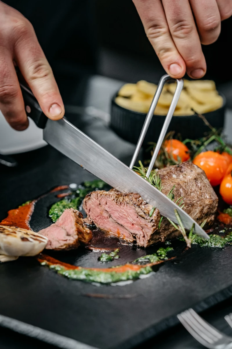 Zartes, rosa gebratenes Rindfleisch wird mit einer Gabel und einem Messer auf einem dunklen Teller angerichtet, umgeben von gebratenen Tomaten, Kräutern und einem Saucenspiegel.