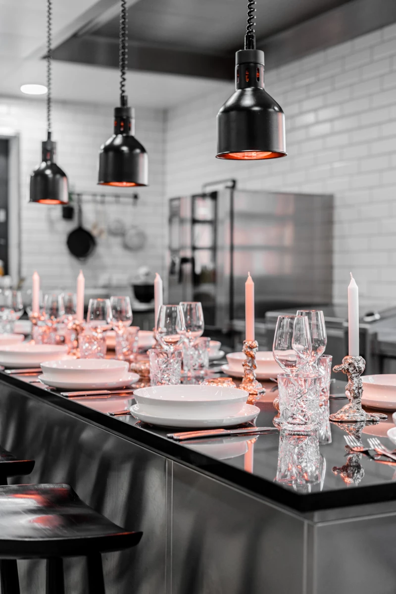 Elegant gedeckter Tisch in moderner Restaurantküche mit schwarzen Hängelampen, weißem Geschirr und Kerzen. Kristallgläser und Dekoration in Pastellrosa auf einer dunklen Theke, die eine stilvolle Tischdekoration betont.