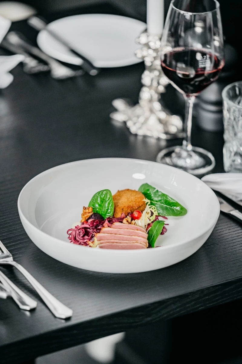 Elegante Fleischplatte mit zartem Schinken, einer orangefarbenen Zitrusfrucht und grünem Basilikum auf einem weißen Teller, daneben ein Glas Rotwein und Silberbesteck auf dunklem Untergrund.