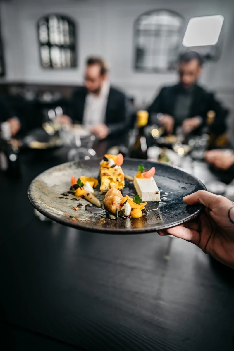 Elegantes Gericht auf einem dunklen Teller mit gelben und orangefarbenen Akzenten, serviert in einem gedämpften Restaurant mit zwei unscharfen Personen im Hintergrund.