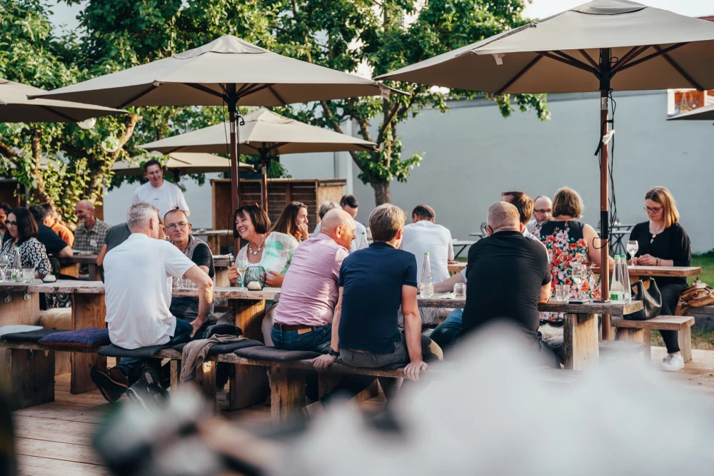 Mehrere Personen sitzen an Holzbänken unter weißen Sonnenschirmen in einem Garten oder auf einer Terrasse. Die Atmosphäre ist entspannt und sommerlich, mit Bäumen im Hintergrund. Verschiedene Altersgruppen unterhalten sich bei einem Sommerfest.