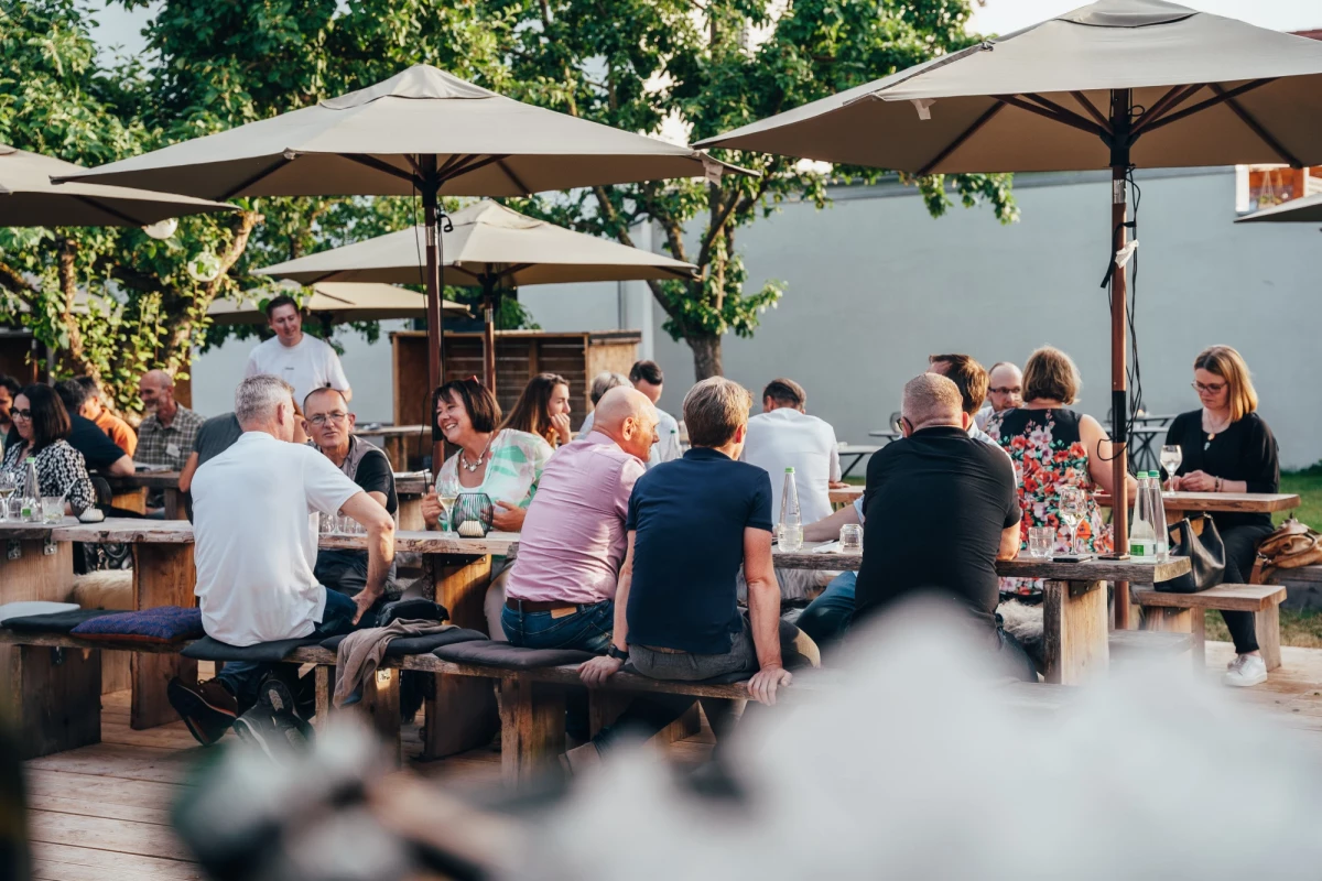 Mehrere Personen sitzen an Holzbänken unter weißen Sonnenschirmen in einem Garten oder auf einer Terrasse. Die Atmosphäre ist entspannt und sommerlich, mit Bäumen im Hintergrund. Verschiedene Altersgruppen unterhalten sich bei einem Sommerfest.
