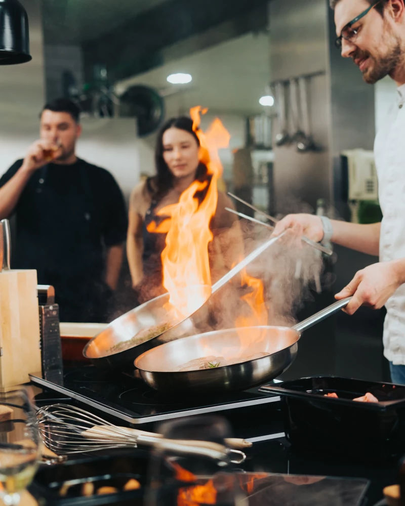 Profikoch flambiert mit hoher Flamme in einer professionellen Restaurantküche. Zwei Kollegen beobachten die spektakuläre Kochszene mit konzentriertem Blick. Kochutensilien und moderne Küchenausstattung sind im Vordergrund sichtbar.