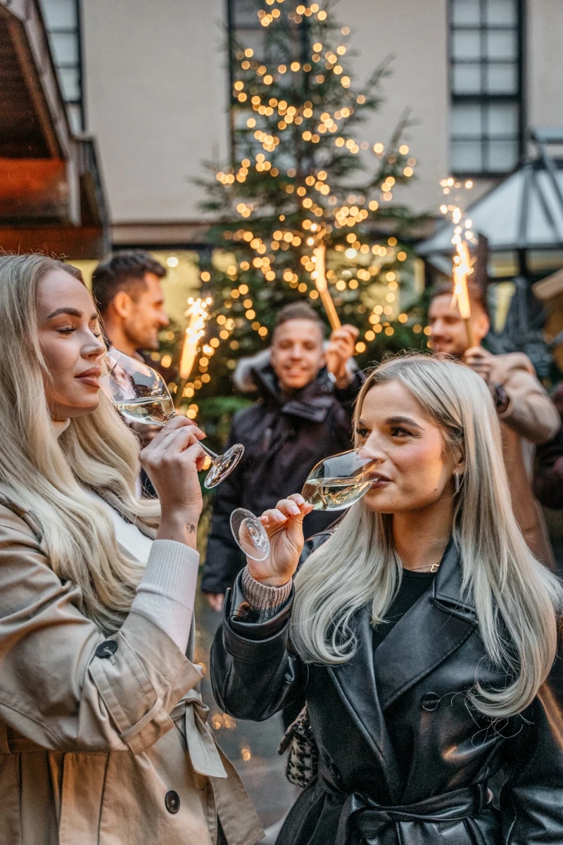 Zwei Frauen mit langen, blonden Haaren trinken Sekt vor einem weihnachtlich beleuchteten Tannenbaum. Sie tragen Wintermäntel und scheinen eine festliche Feiertagsstimmung zu genießen. Im Hintergrund sind weitere Personen in einer gemütlichen, weihnachtlichen Atmosphäre zu sehen.