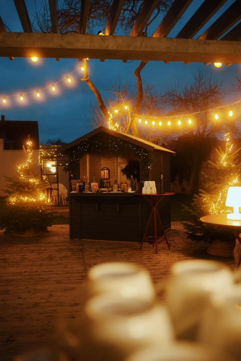 Beleuchteter Weihnachtsmarktstand mit warmen Lichterketten in einer gemütlichen Winterabendatmosphäre. Holzpergola und Verkaufsstand sind in goldene Lichter getaucht, umgeben von Weihnachtsbäumen und einer stimmungsvollen Abendkulisse.
