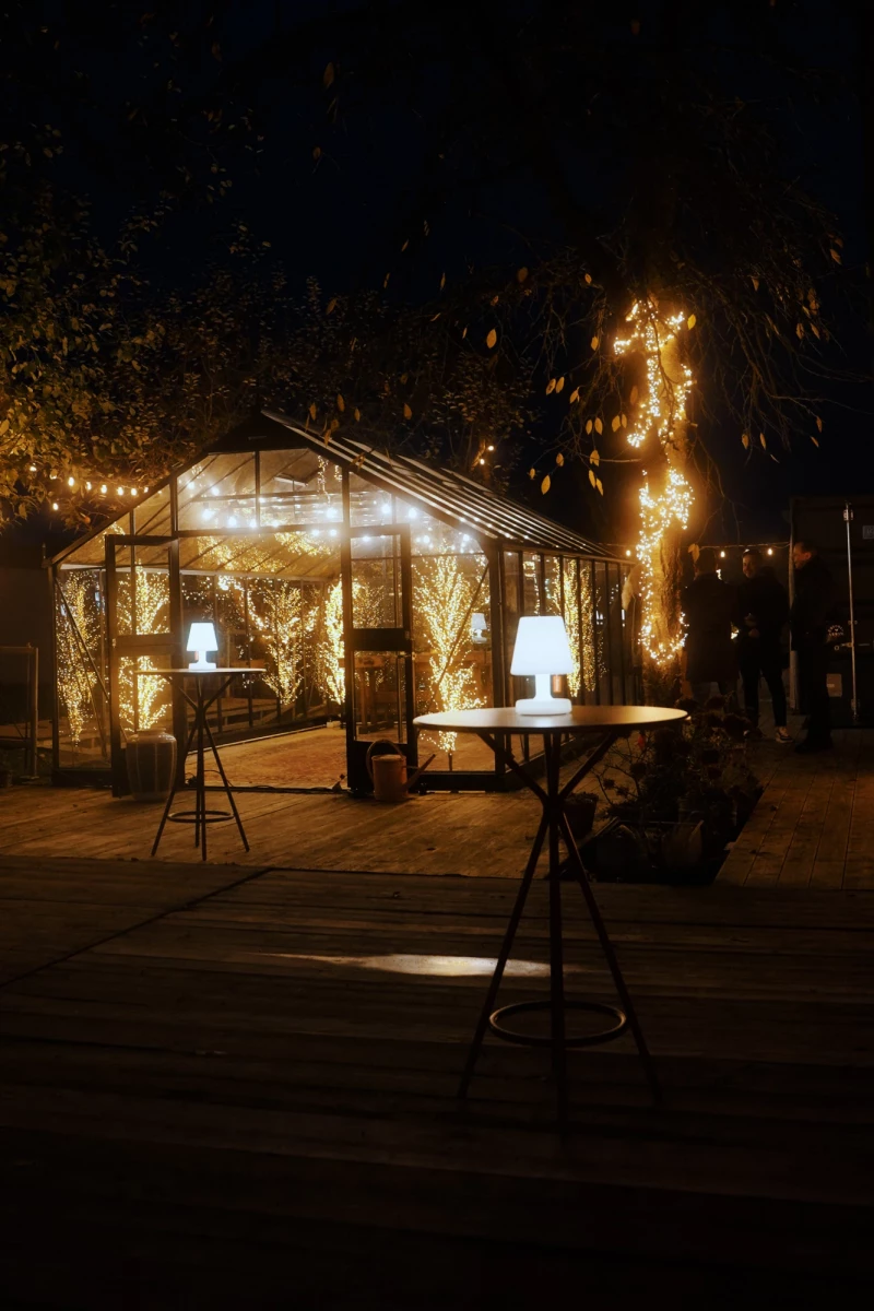 Beleuchtetes Gewächshaus in der Nacht mit warmen Lichterketten, umgeben von dunklen Bäumen. Zwei Stehlampen und Tische im Vordergrund auf einer Holzterrasse, die eine gemütliche und romantische Atmosphäre ausstrahlen.
