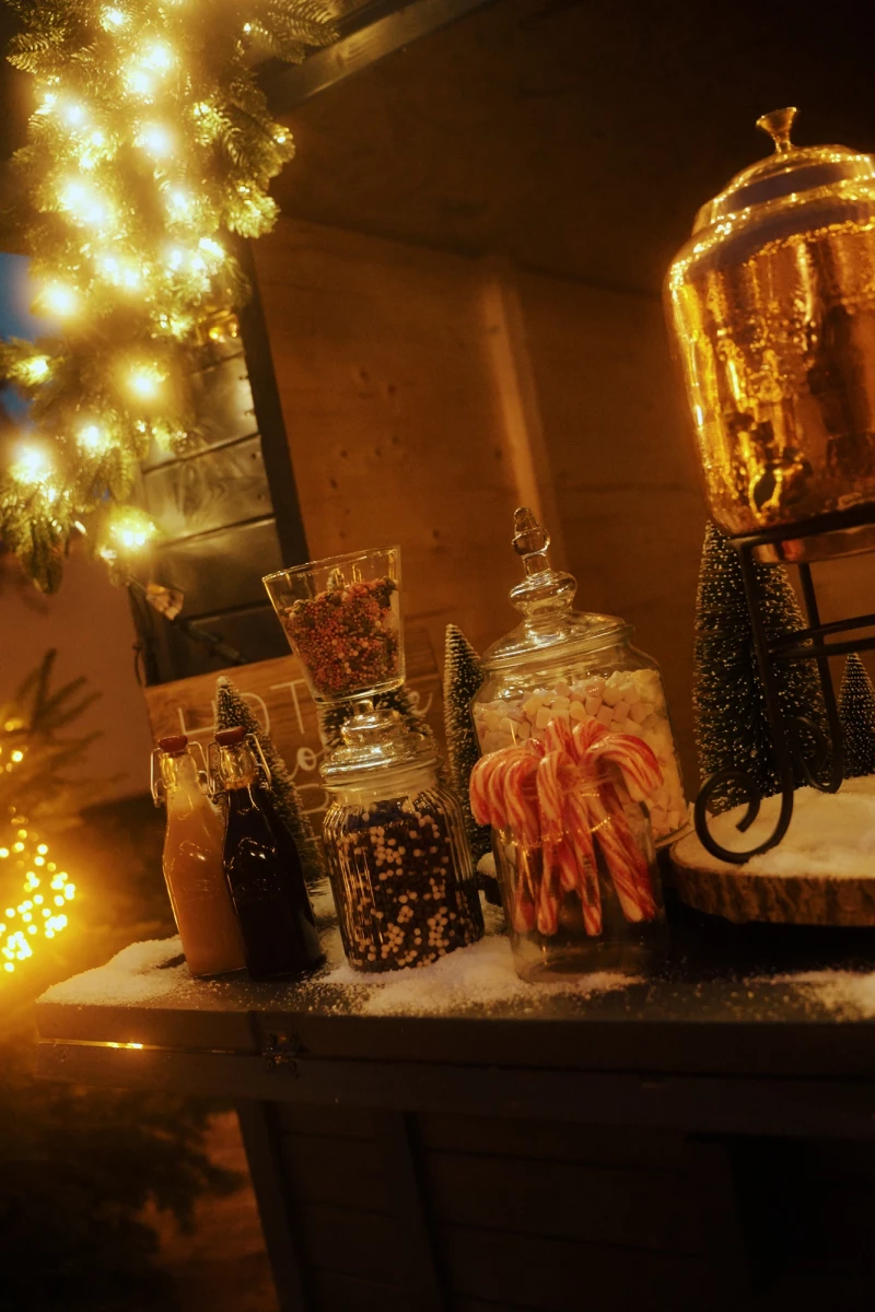 Weihnachtlich beleuchtete Theke mit Glasgefäßen voller Kaffee, Süßigkeiten und Zimtstangen, im Hintergrund ein beleuchteter Weihnachtsbaum in warmen, goldenen Tönen.