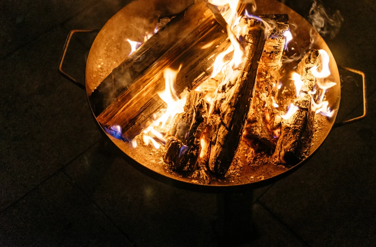 Loderndes Lagerfeuer in einer Feuerschale mit brennenden Holzscheiten, die hell und intensiv in der Dunkelheit leuchten. Die Flammen tanzen um die Holzstücke und erzeugen eine warme, einladende Atmosphäre.
