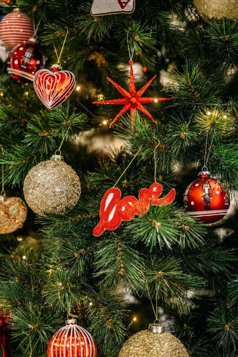 Nahaufnahme eines Weihnachtsbaums mit verschiedenen Christbaumkugeln in Gold und Rot, darunter eine herzförmige Kugel, eine Schneeflocke und eine rote Schriftzug "Love". Die Kugeln hängen an den grünen Zweigen des Tannenbaums.