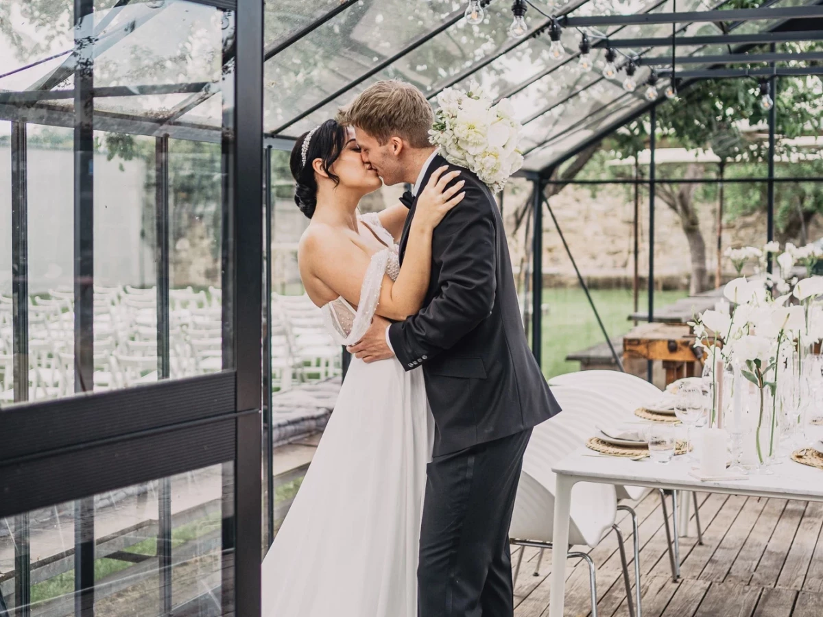 Brautpaar in einem lichtdurchfluteten Gew&auml;chshaus, eng umarmend und k&uuml;ssend auf einem Holzdeck. Die Szene ist minimalistisch und elegant gestaltet, mit wei&szlig;en Kerzen und gedecktem Tisch im Hintergrund, umrahmt von Glasw&auml;nden und Metallrahmen.