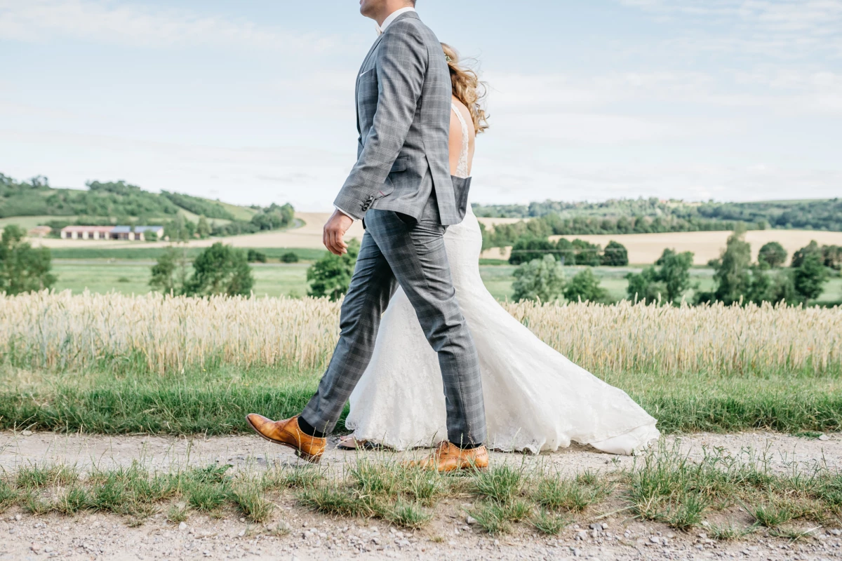 Brautpaar auf einem Feldweg, der Bräutigam in grauem Anzug und gelben Schuhen, die Braut in einem weißen Brautkleid, im Hintergrund eine weite Landschaft mit Feldern und Bäumen.