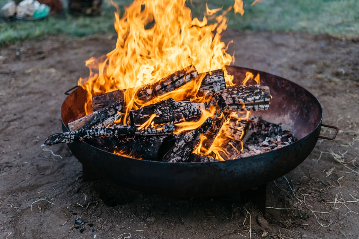 Brennende Holzscheite in einer schwarzen Feuerschale auf dem Boden. Lodernde Flammen in Gelb- und Orangetönen umgeben die Holzkohle. Die Glut leuchtet hell vor einem unscharfen Hintergrund.