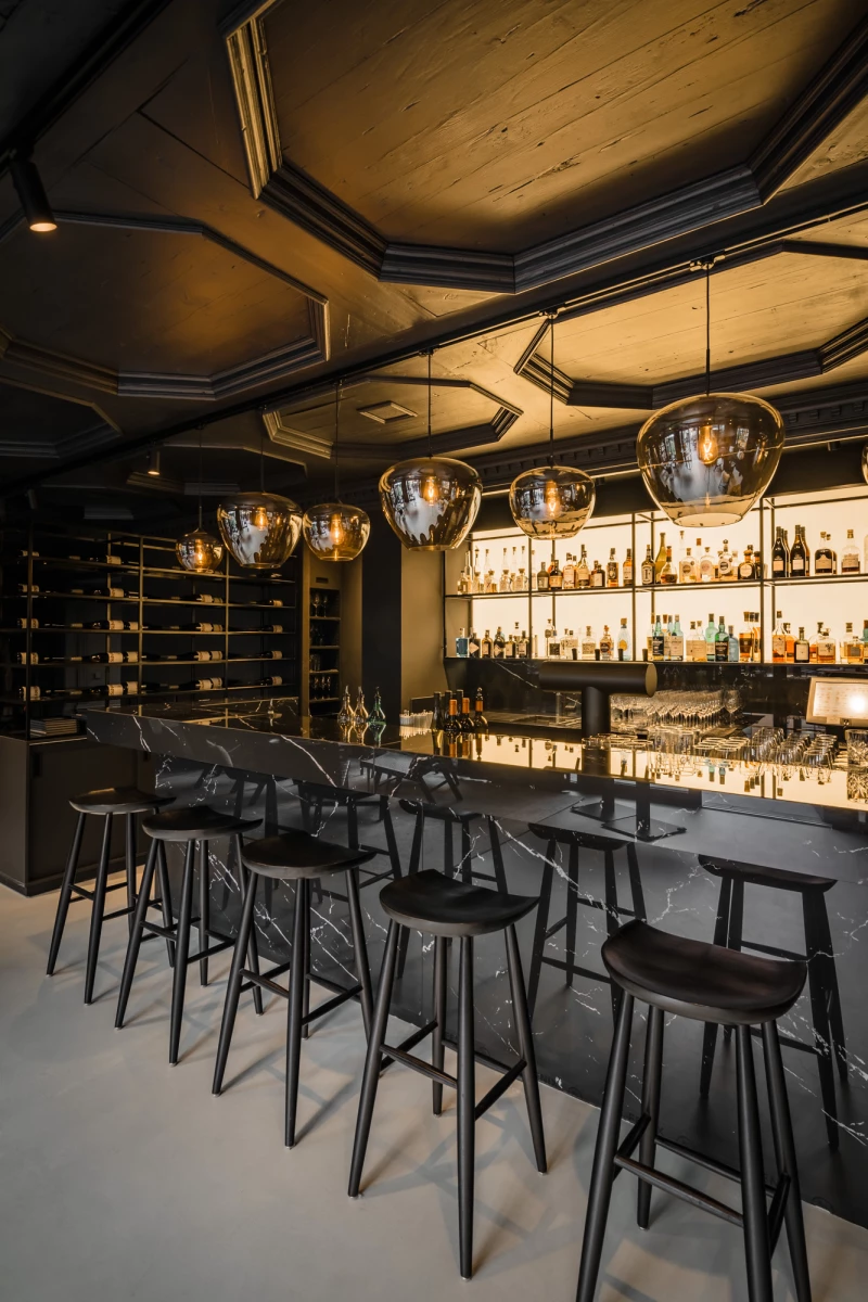 Elegante Bar mit dunklen Holzhockern, die an einer langen Theke stehen. Über der Theke hängen große, runde Pendelleuchten, und die Rückwand ist mit beleuchteten Regalen voller Spirituosenflaschen bestückt. Die gedämpfte Beleuchtung und die warmen Holztöne schaffen eine einladende, edle Atmosphäre.
