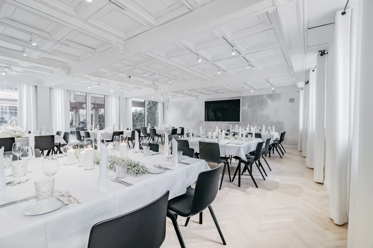 Eleganter Veranstaltungsraum in Weiß und Schwarz mit gedeckten Tischen, schwarzen Stühlen und einem großen Fernseher an der Wand. Die Decke ist mit Spotlights ausgestattet und große Fenster lassen viel natürliches Licht herein, was eine helle und moderne Atmosphäre schafft.