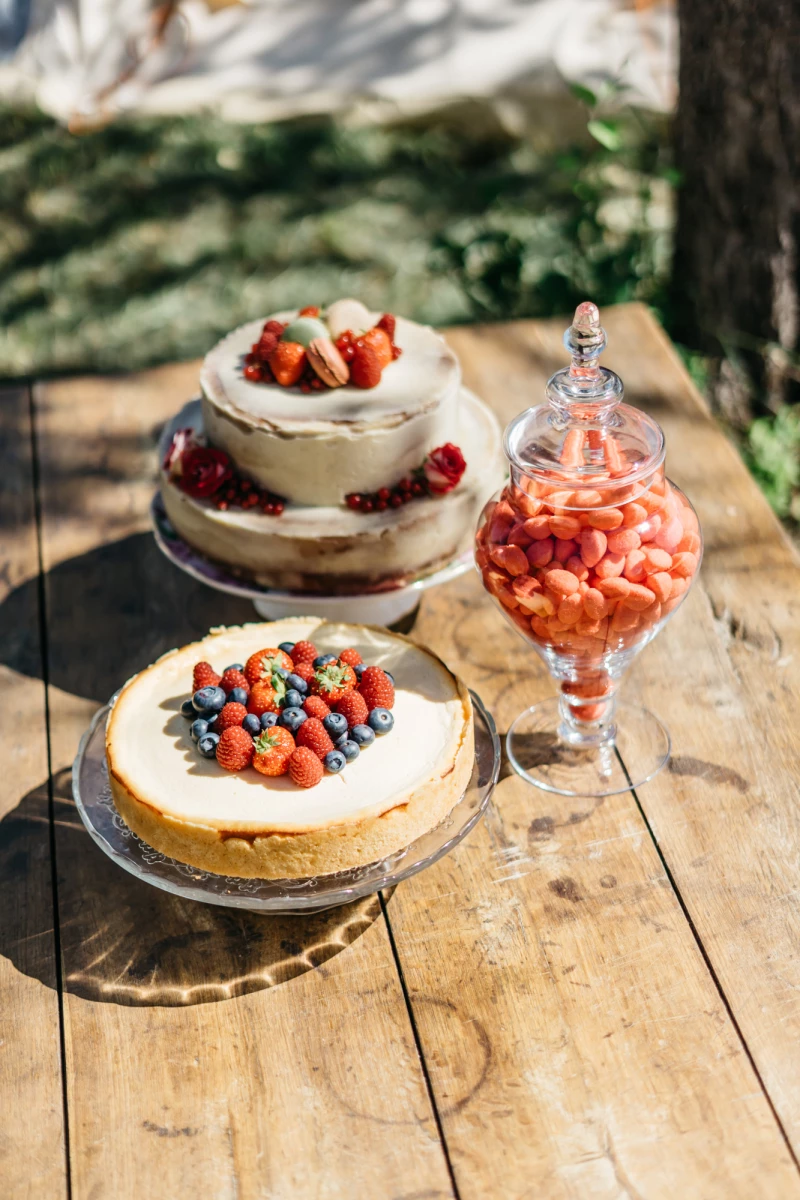 Zwei Obsttorten mit Beeren auf einem rustikalen Holztisch, daneben ein Glas mit roten Erdbeeren. Die Torten sind mit verschiedenen Beerensorten wie Erdbeeren, Blaubeeren und Himbeeren dekoriert, vor einem unscharfen grünen Hintergrund.