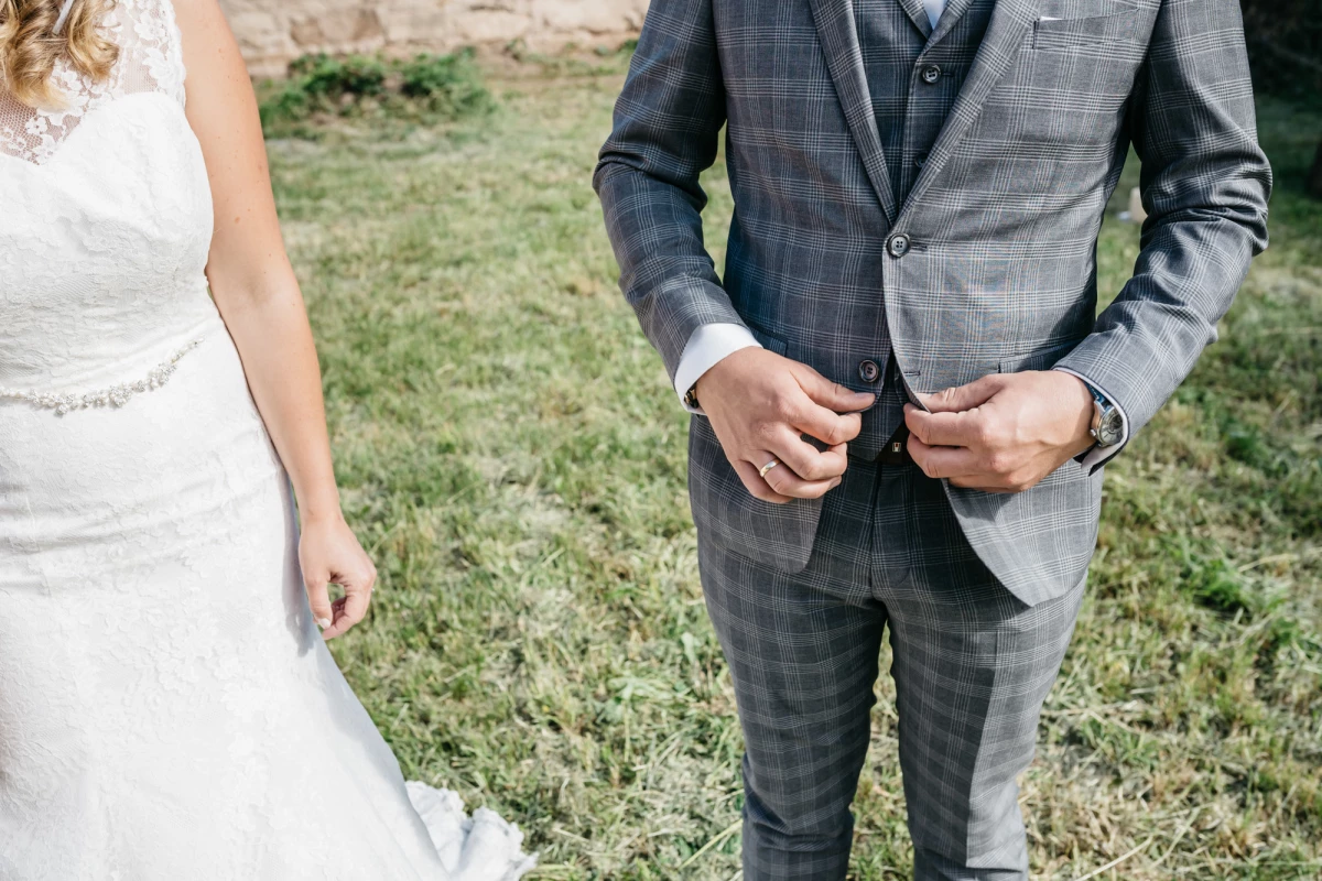 Detailaufnahme einer Hochzeitsszene mit einem Bräutigam in einem grauen karierten Anzug, der seine Jacke schließt, neben einer Braut in einem weißen Spitzenkleid auf einer Wiese.