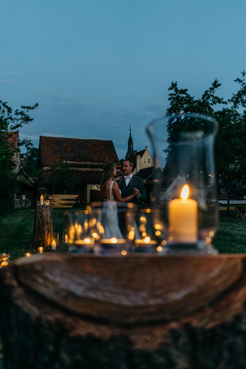 Abenddämmerung auf einem romantischen Hochzeitsort mit Kerzen im Vordergrund und verschwommenen Brautleuten im Hintergrund. Rustikale Holzscheibe und brennende Kerzen bilden den Rahmen für eine stimmungsvolle Szene mit einer Kirche und Bäumen in der Dämmerung.
