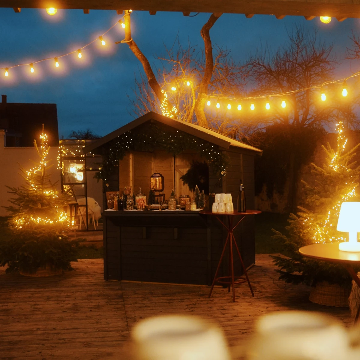 Weihnachtlich beleuchtete Außenbar mit warmweißen Lichterketten, umgeben von verschneiten Bäumen und einer gemütlichen Holzhütte im Hintergrund, die eine festliche Winterstimmung an einem dunklen Abend einfängt.