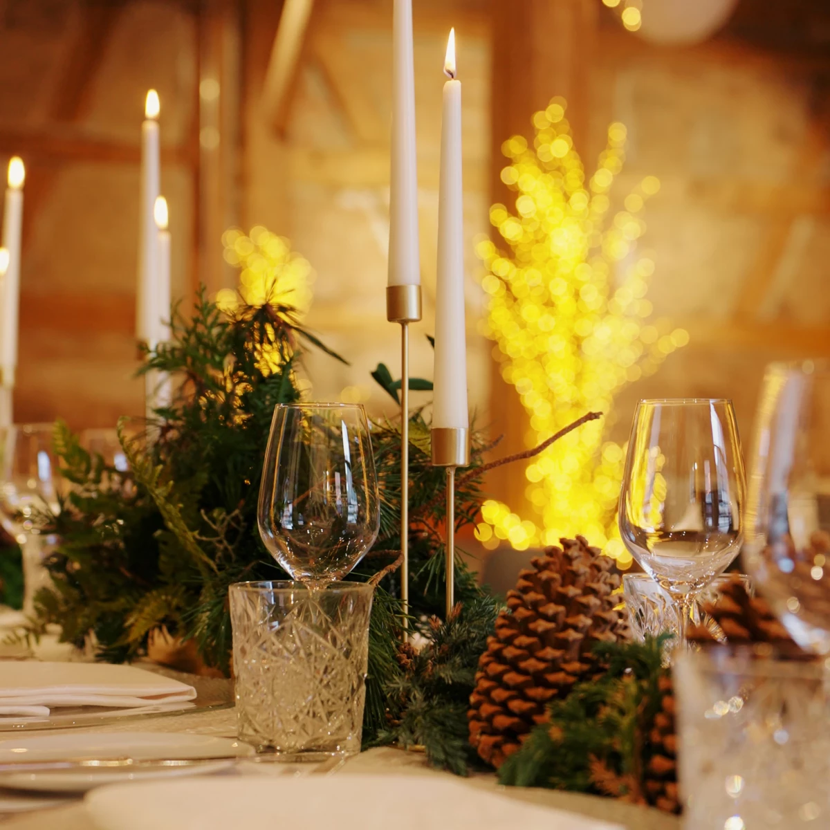 Festlich gedeckter Tisch mit weißen Kerzen in Messinghaltern, Weingläsern und Tannenzweigen, dazu große Tannenzapfen. Im Hintergrund verschwommene gelbe Lichter, die eine warme, weihnachtliche Atmosphäre erzeugen.
