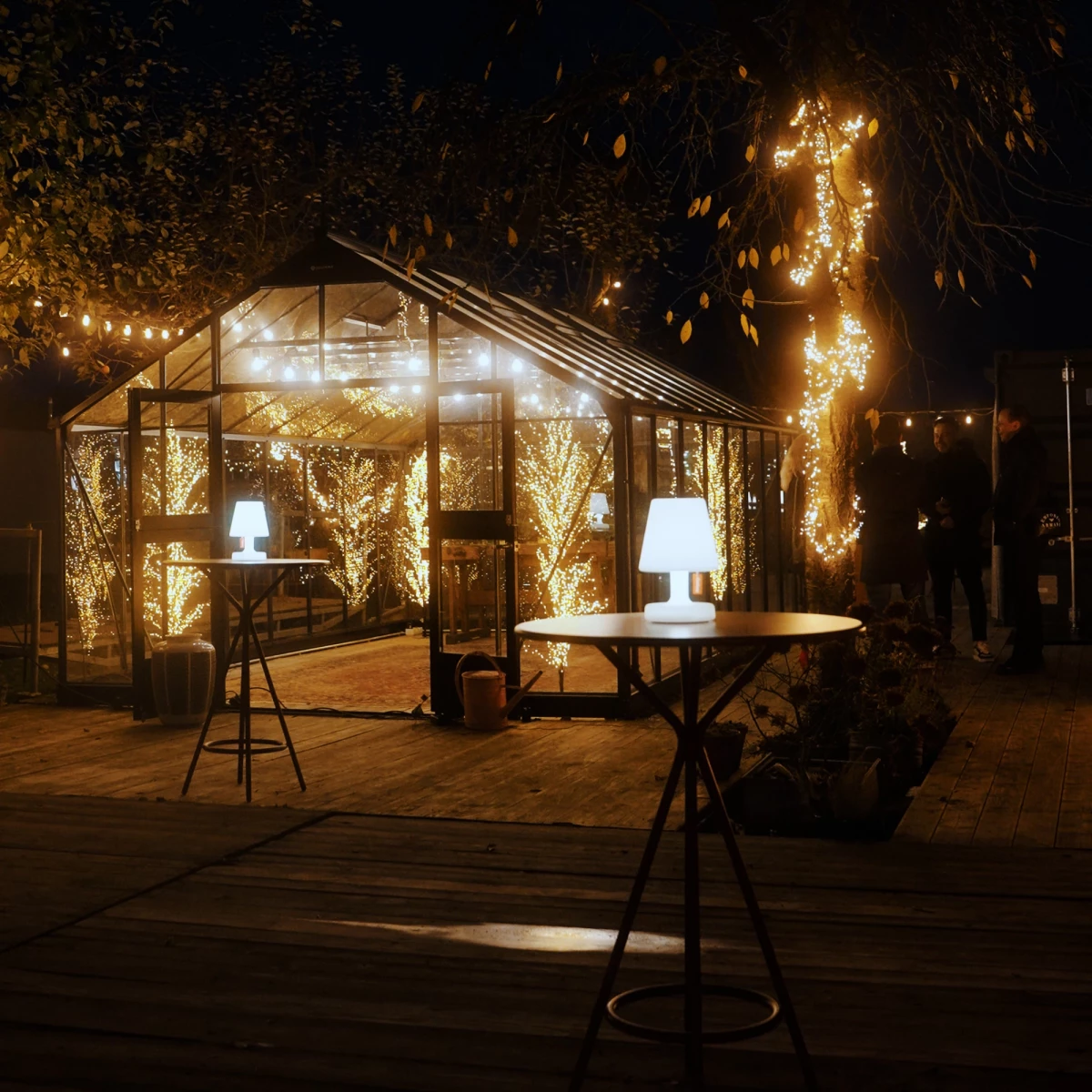 Beleuchtetes Gewächshaus in der Nacht mit warmen Lichterketten, umgeben von dunklen Bäumen. Zwei Stehlampen und Tische im Vordergrund auf einer Holzterrasse, die eine gemütliche und romantische Atmosphäre ausstrahlen.