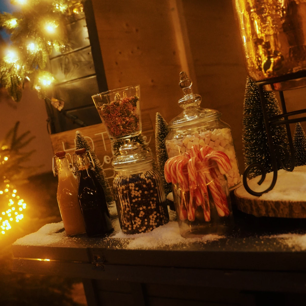 Weihnachtliche Szenerie mit Glasgefäßen voller Süßigkeiten, Lichterketten und Weihnachtsdekoration, die auf schneebedecktem Untergrund in warmem, goldenem Licht glänzen.