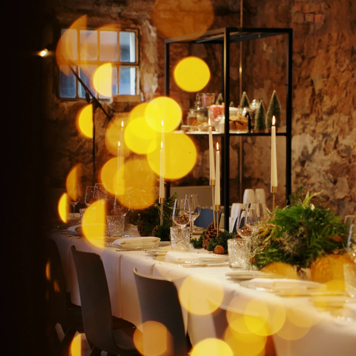 Festlich gedeckter Tisch in rustikaler Steinumgebung mit weißer Tischdecke, Gründekorationen und gelben Lichtreflexen. Weingläser und Kerzen auf dem Tisch, im Hintergrund ein Regalgestell und ein Fenster, das warme Atmosphäre vermittelt.