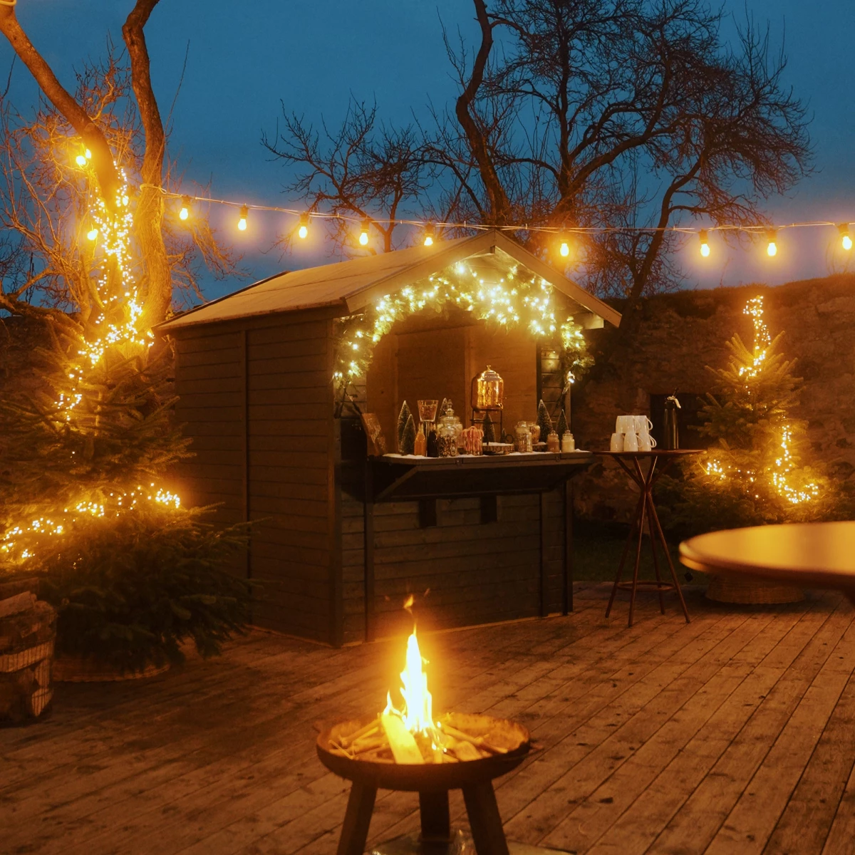Romantische Winterabendszene mit Lagerfeuer auf einer Holzterrasse, umgeben von beleuchteten Bäumen und Lichterketten, mit einer gemütlichen Holzhütte im Hintergrund, die weihnachtlich geschmückt ist und eine warme, einladende Atmosphäre ausstrahlt.