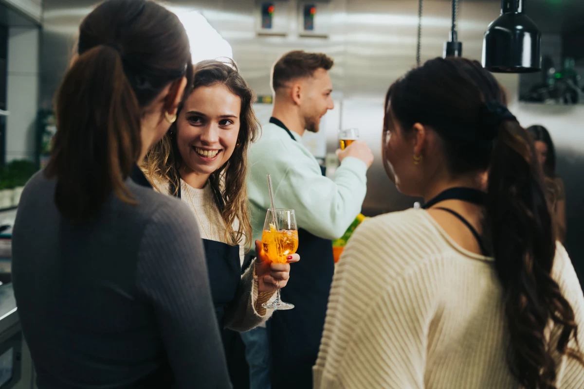 Drei junge Frauen unterhalten sich bei einer Feier in einem Raum, eine lächelt und hält ein orangefarbenes Getränk. Im Hintergrund steht eine weitere Person, die sich abwendet.