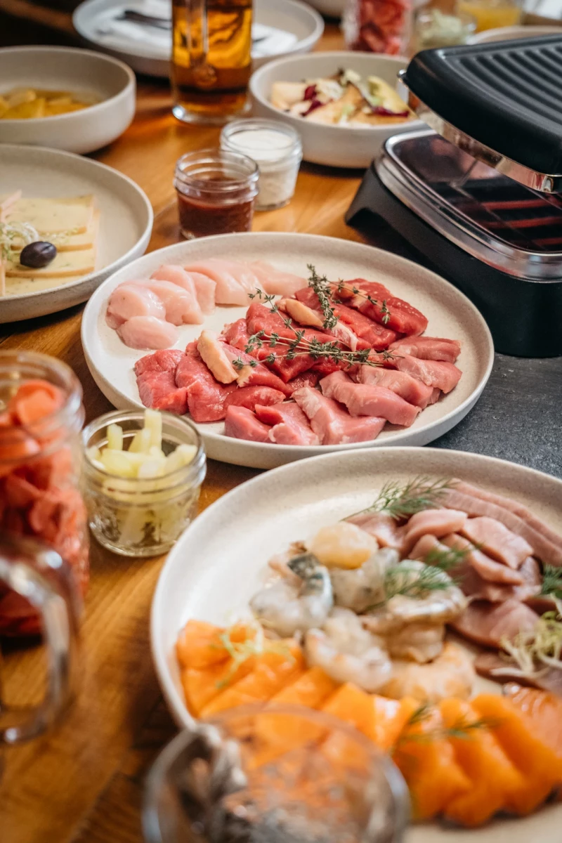Verschiedene Teller mit Aufschnitt, Fleisch und Wurstwaren in Pastellfarben, dekoriert mit Kräutern. Ein Raclette- oder Fondue-Setup mit verschiedenen Fleischsorten und Beilagen auf einem Holztisch.