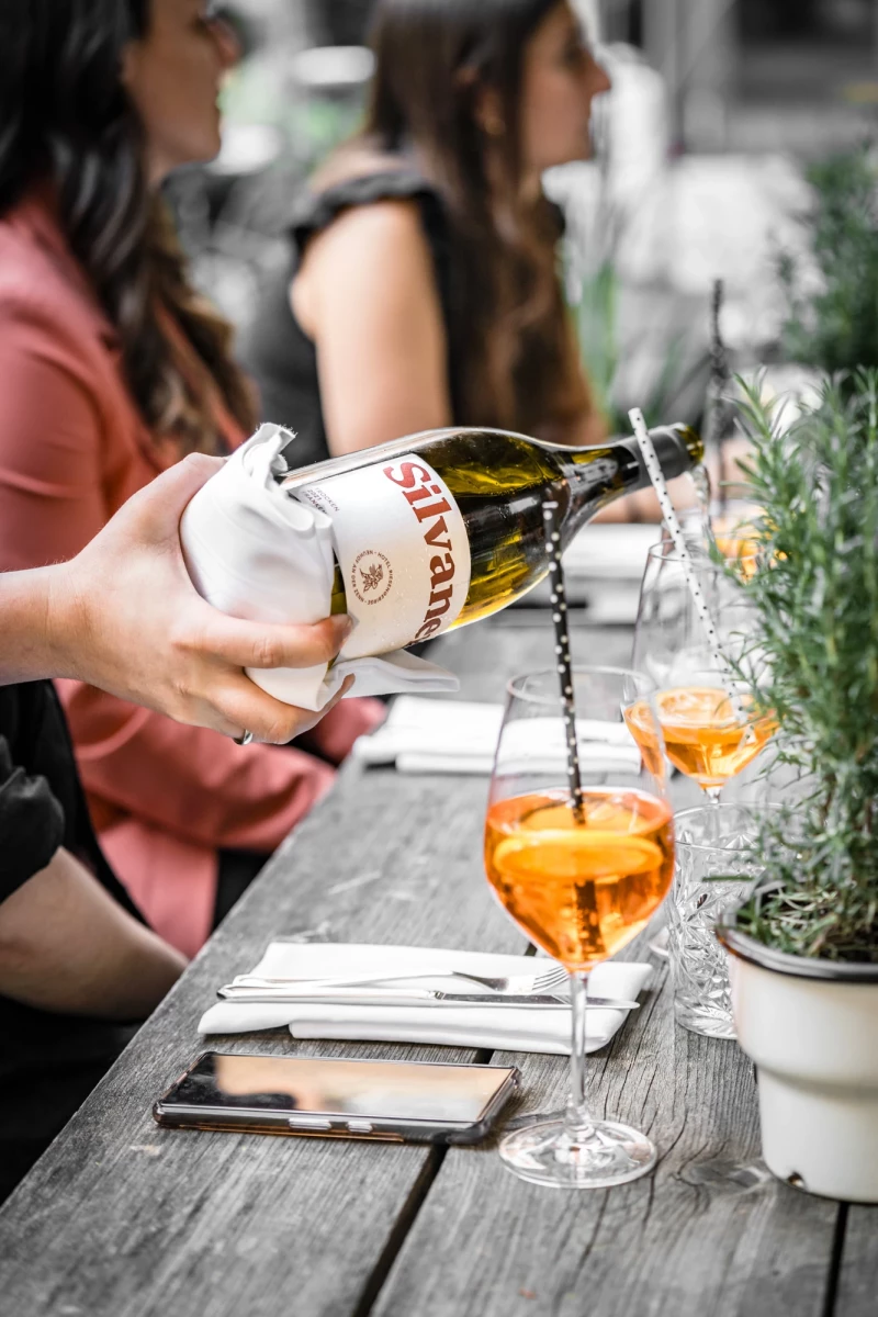 Hand gießt einen orangefarbenen Cocktail aus einer Flasche in ein Weinglas auf einem grauen Holztisch, im Hintergrund sitzen Menschen an einem Tisch mit Rosmarin-Pflanze.