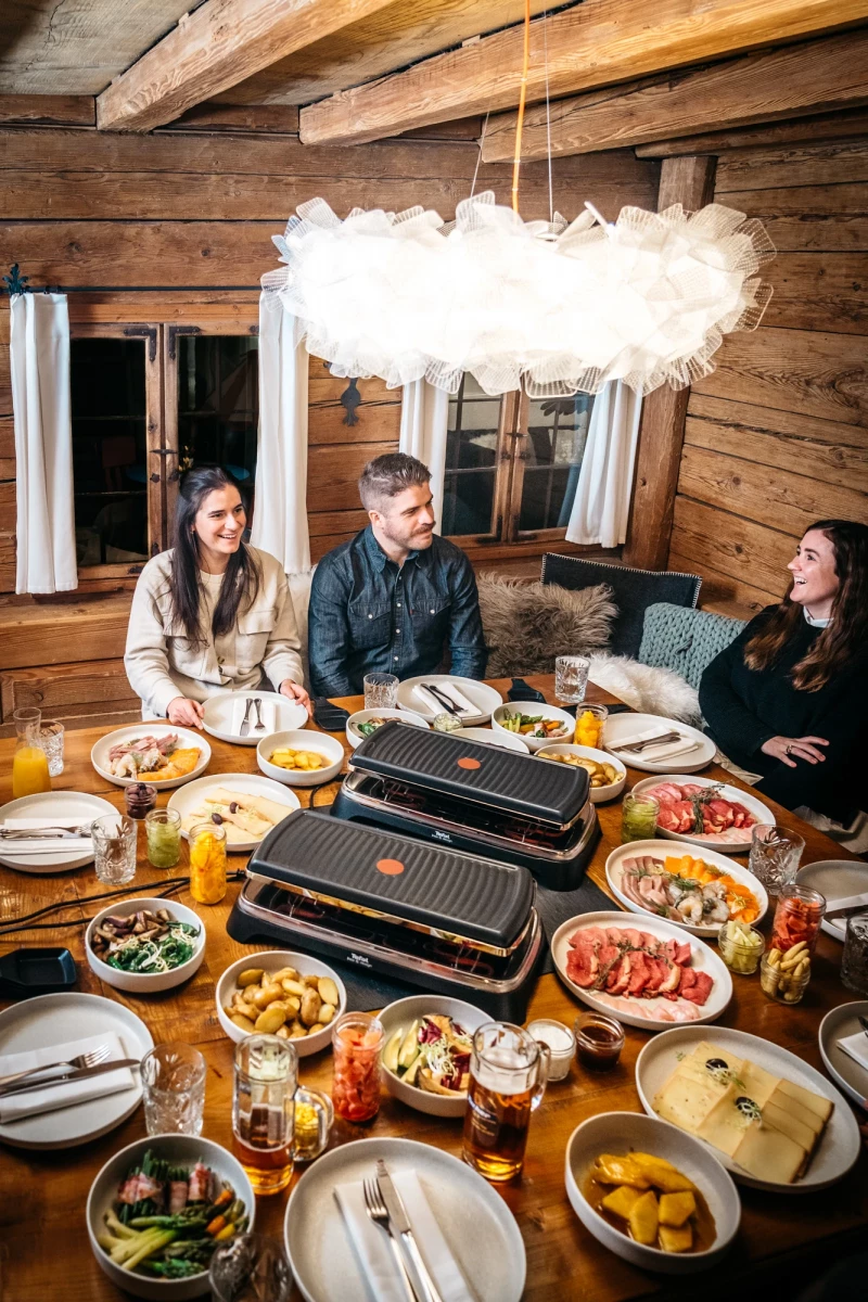 Drei Personen sitzen an einem Holztisch mit reichlich gedeckter Raclette-Grillplatte und verschiedenen Zutaten. Eine weiche Wolkenlampe hängt über dem gemütlichen Holztisch in einer rustikalen Holzhütte, während die Gruppe bereit ist, gemeinsam zu essen.