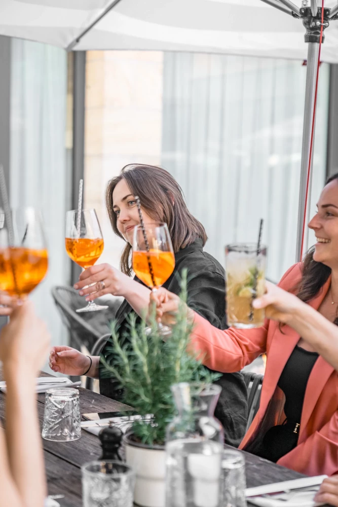 Zwei Frauen stoßen mit orangefarbenen Cocktails in einem hellen Innenraum an. Sie tragen schwarze und lachsfarbene Kleidung und sitzen an einem Tisch mit Pflanzendekoration. Die Stimmung ist fröhlich und entspannt.