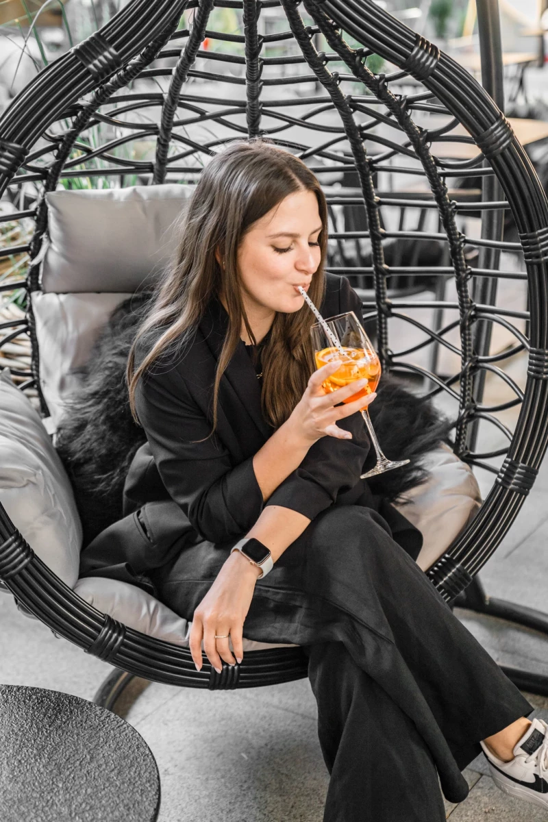 Eine Frau in schwarzer Kleidung sitzt in einem schwarzen, geschwungenen Hängesessel und hält ein orangefarbenes Cocktailglas. Sie trägt eine Smartwatch und scheint entspannt in einer gemütlichen Loungeumgebung zu sein.