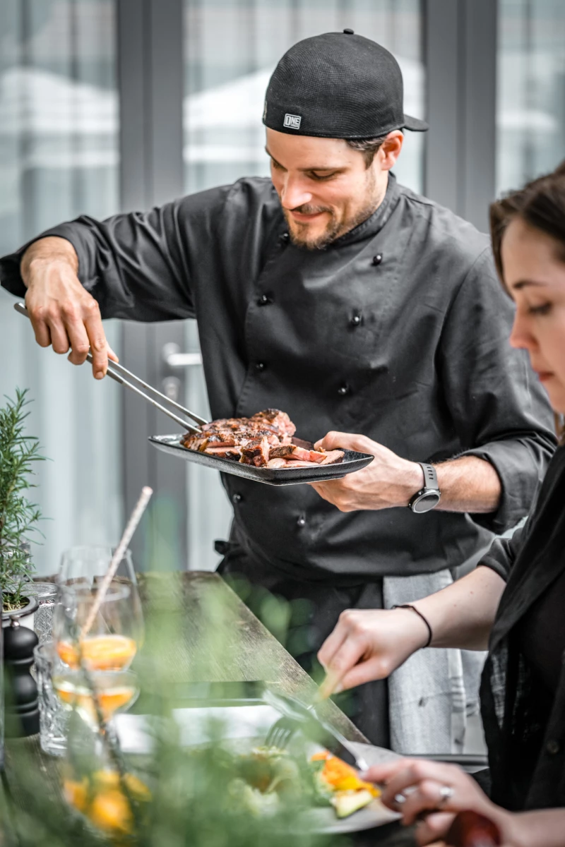 Koch in schwarzer Uniform und Mütze serviert gebratenes Fleisch von einem Teller, während eine Kollegin im Hintergrund mit einer Zitrone hantiert. Professionelle Küchensituation mit appetitlich zubereiteten Speisen.