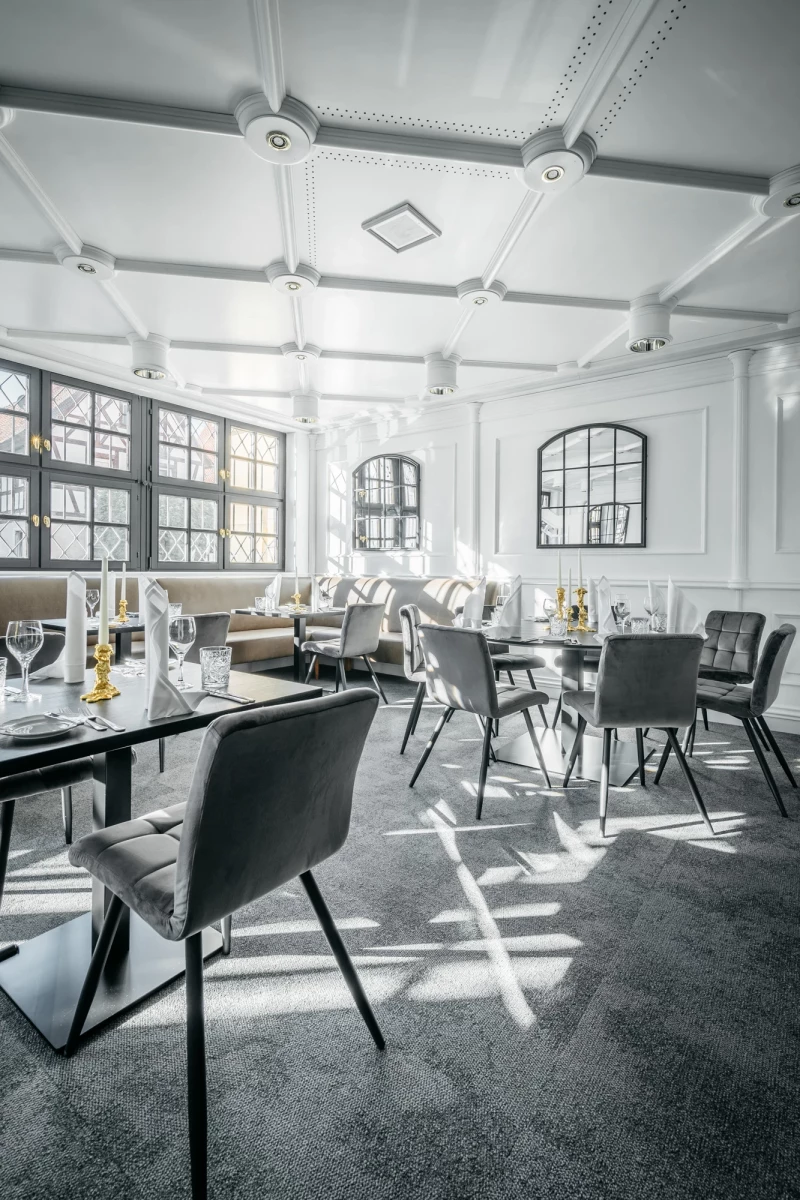 Moderner, lichtdurchfluteter Restaurantbereich mit weißen Wänden, großen Fenstern und grauen Designerstühlen. Elegante Tische sind gedeckt, und der Raum strahlt eine helle, luftige Atmosphäre aus.