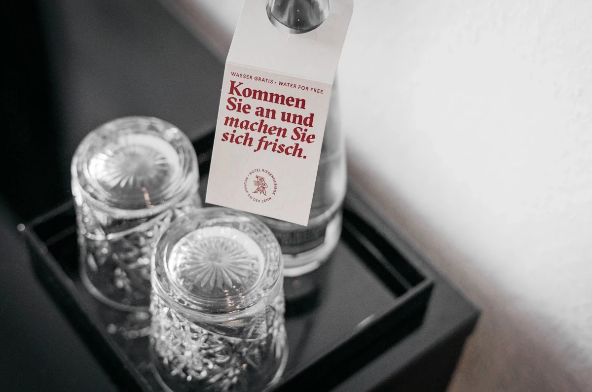 2 umgedrehte Gläser und eine Wasserflasche stehen auf einem dunklen Tablett. An der Wasserflasche hängt ein Papier-Anhänger mit der Aufschrift "Wasser gratis. Kommen Sie an und machen Sie sich frisch."