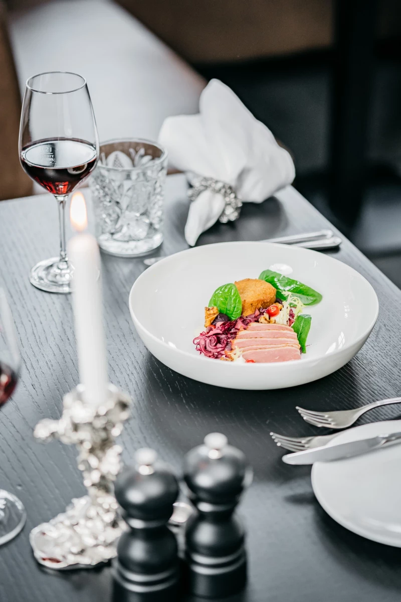Elegantes Restaurantgedeck mit einem weißen Teller, auf dem ein künstlerisch angerichtetes Gericht mit Rosa-Tönen, grünem Blattwerk und einer orangefarbenen Komponente serviert wird. Ein Glas Rotwein und eine brennende Kerze vervollständigen die stilvolle Tischdekoration.