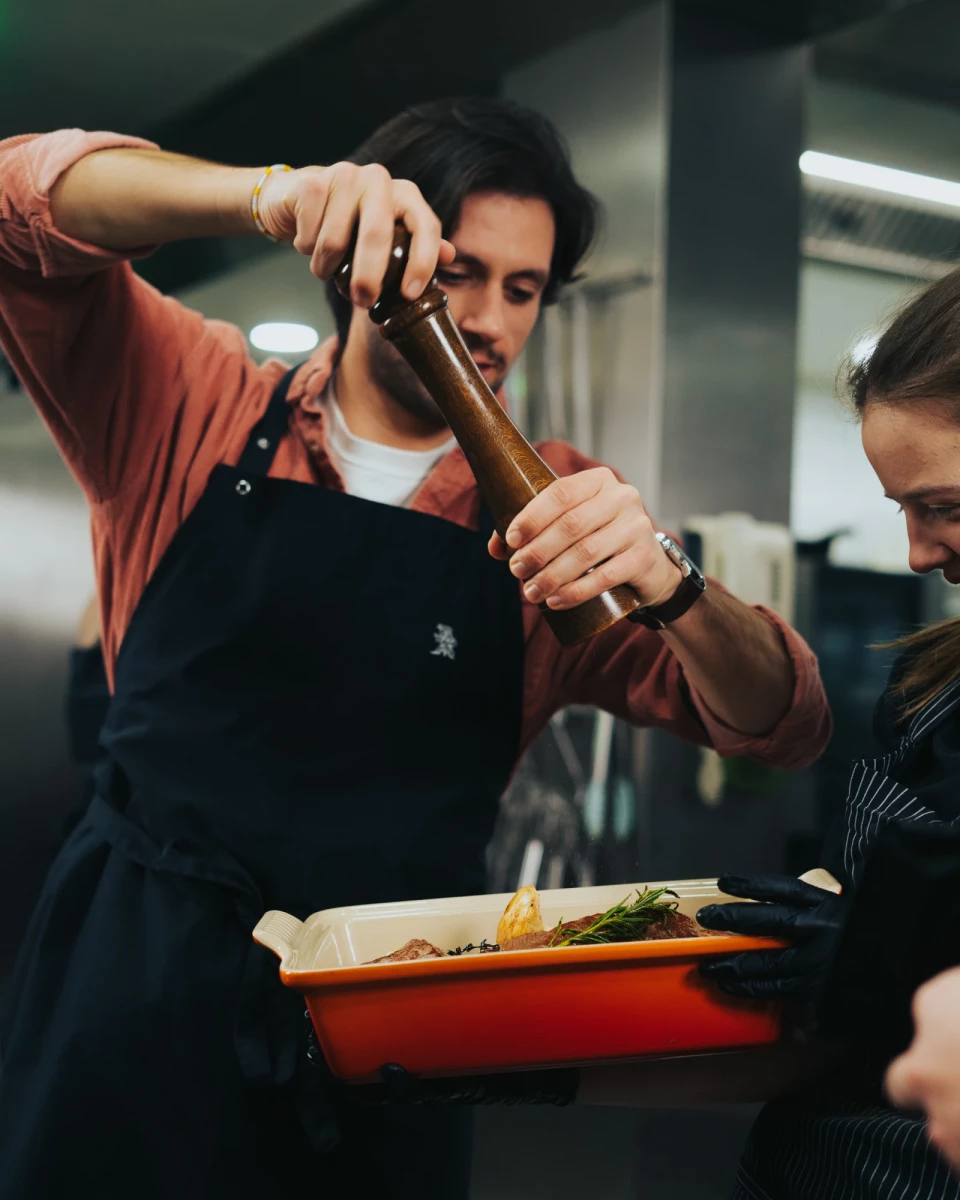 Koch in schwarzer Schürze benutzt einen Stampfer oder Stößel, um eine Zutat auf einem orangefarbenen Tablett zuzubereiten. Ein anderer Koch steht daneben und beobachtet die Zubereitung in einer professionellen Restaurantküche.