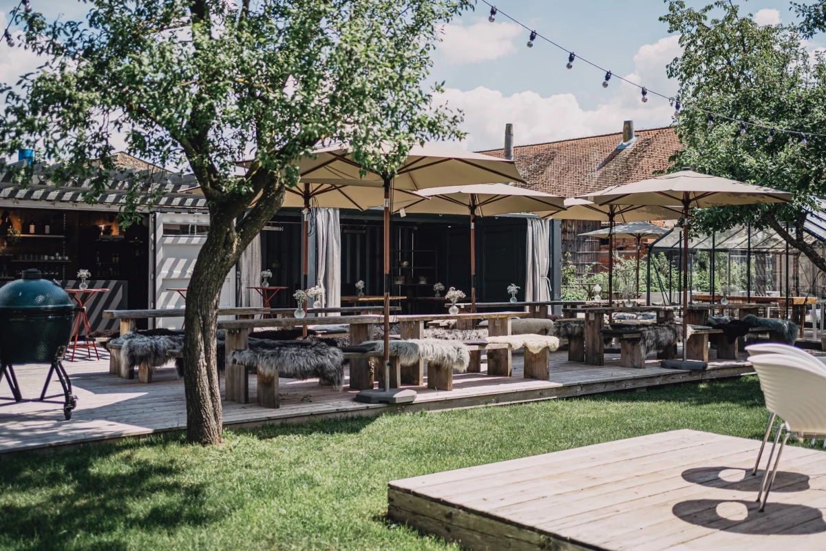 Sonnige Terrasse mit weißen Sonnenschirmen, Holzmöbeln und einem Baum, umgeben von gepflegtem Rasen und rustikalen Gebäuden im Hintergrund, perfekt für entspannte Mahlzeiten im Freien.