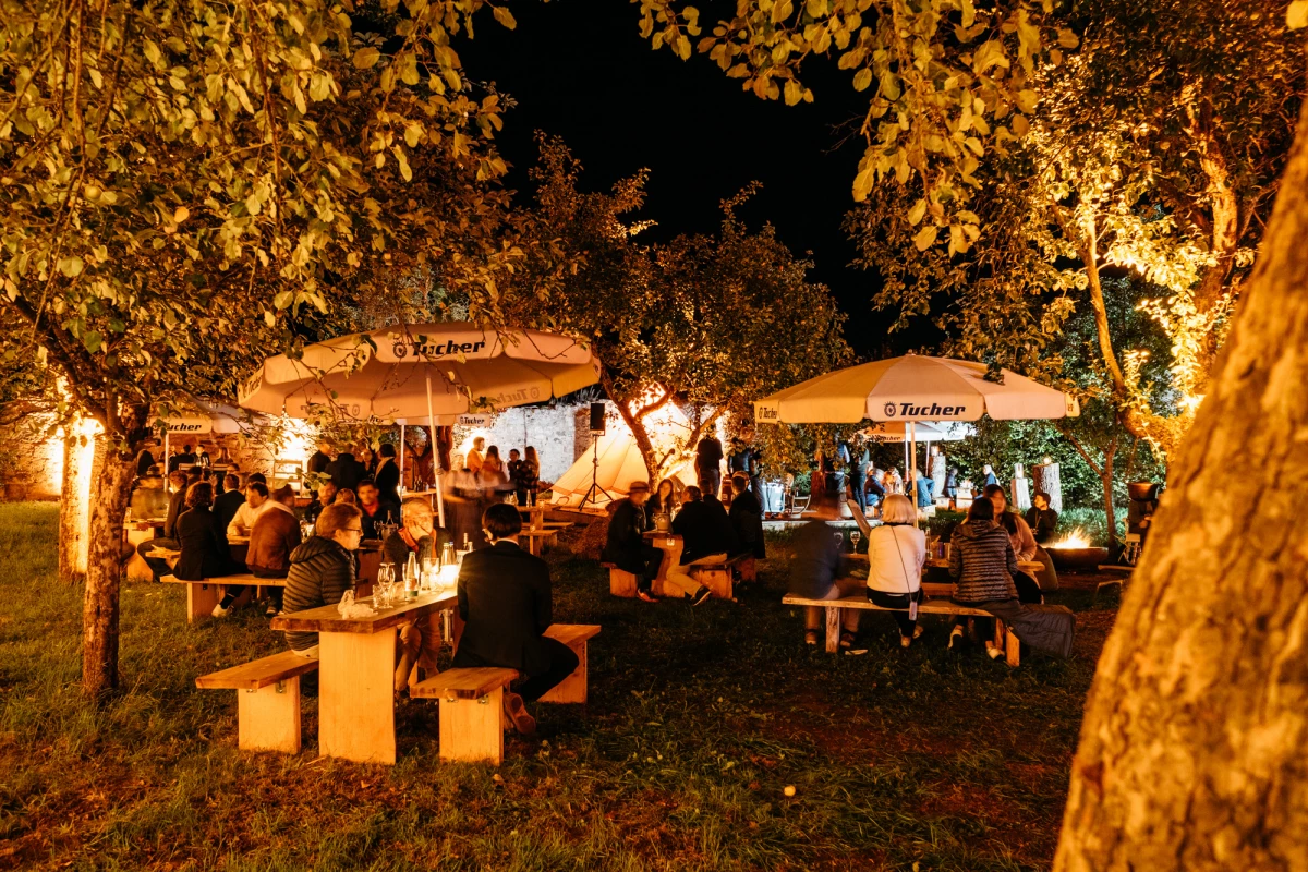 Abendliche Gartenszene mit beleuchteten Pavillons und Stehtischen, Menschen versammeln sich unter herbstlichen Bäumen mit warmgoldenem Licht und gedämpfter Atmosphäre.