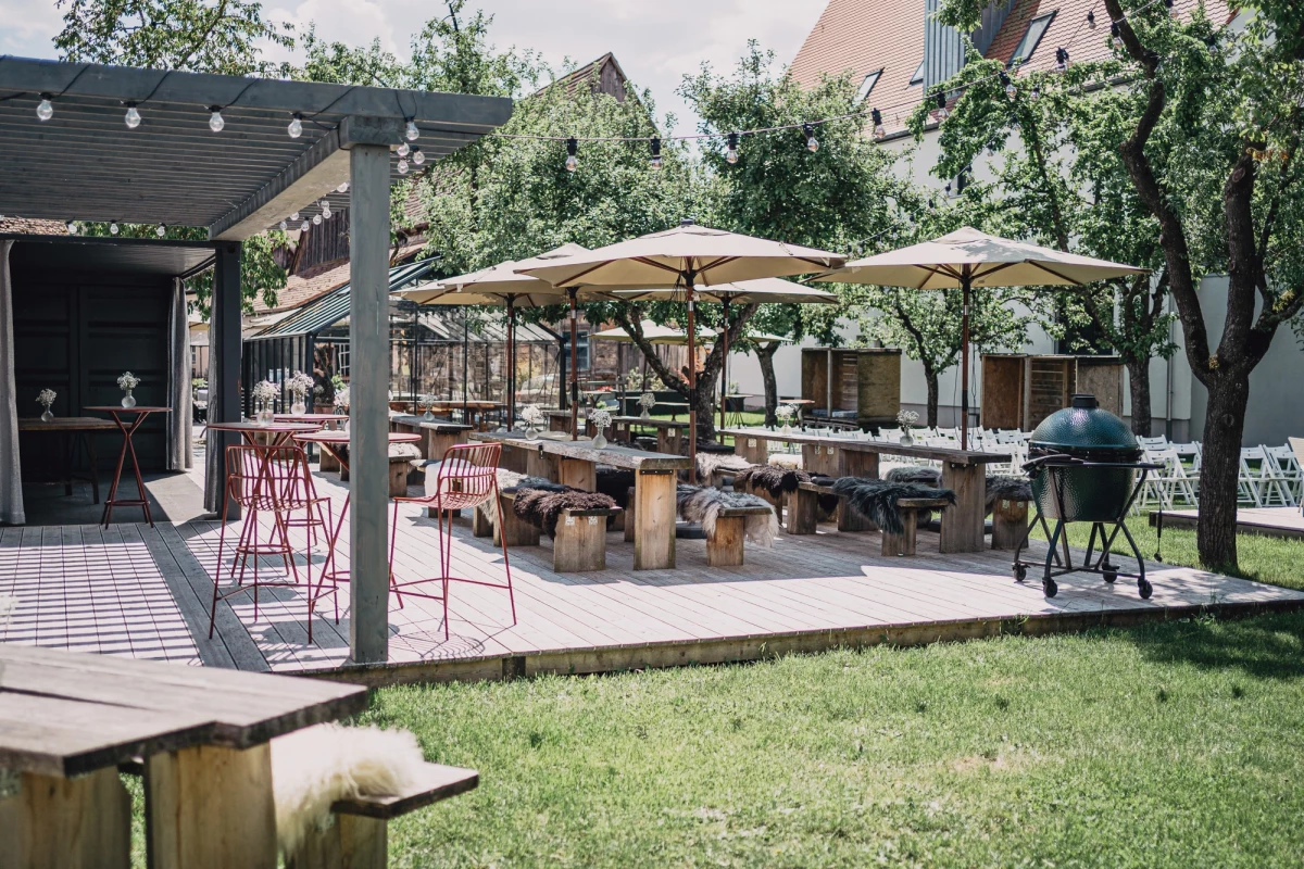 Sonnige Gartenterrasse eines Hotels mit Holztischen, weißen Sonnenschirmen und einer überdachten Sitzfläche. Grüne Rasenfläche und Bäume rahmen den gemütlichen Außenbereich ein, der zum Verweilen einlädt.