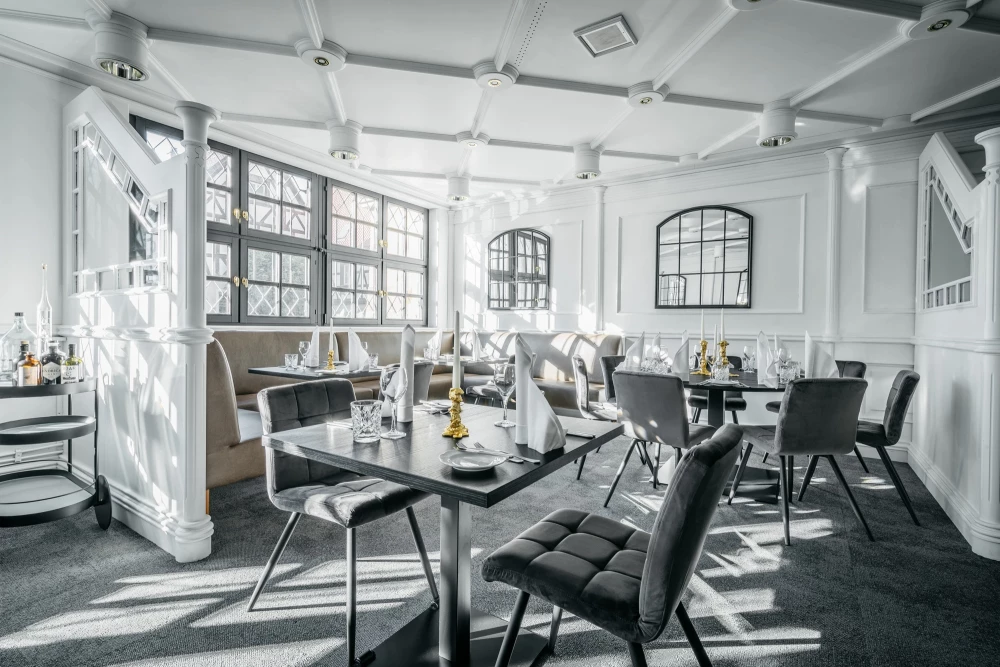 Helles, luftiges Restaurantinterieur mit modern designten Stühlen und gedeckten Tischen. Große Fenster lassen viel natürliches Licht herein, und weiße Wände sowie Säulen verleihen dem Raum eine elegante, offene Atmosphäre.