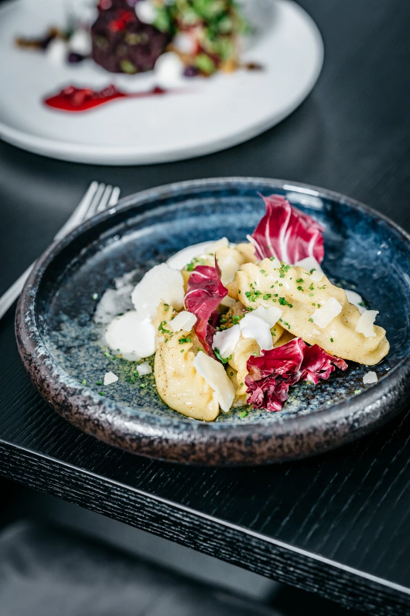 Feine Kartoffelgnocchi auf einem blauen Teller, garniert mit rosa Radicchio-Blättern und einer cremigen Sauce. Die Speise ist kunstvoll angerichtet und liegt auf einem dunklen, strukturierten Untergrund.